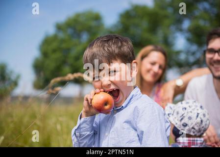 Kleiner Junge, der einen Apfel auf der Wiese isst, auf dem Land, mit Familie im Hintergrund, Bayern, Deutschland Stockfoto