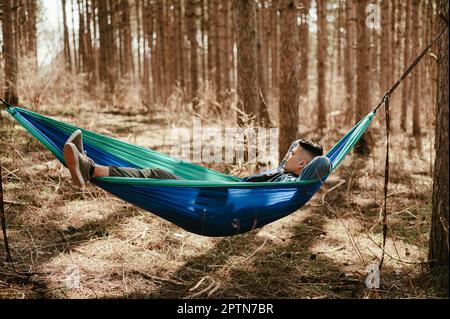 Junger Mann, der in einer blauen Hängematte im Pine Tree Forest liegt oder sich ausruht Stockfoto