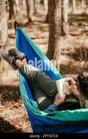 Junger Mann, der sich in der Hängematte entspannt und in einem Journal im Wald schreibt Stockfoto