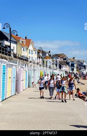 Touristen laufen entlang der Promenade mit dem Strand auf der rechten Seite und Strandhütten auf der linken Seite, Lyme Regis, Dorset, Großbritannien. Stockfoto