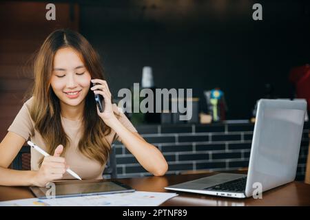 Lächelnde Frau, die mit einem Laptop arbeitet, und notieren Sie sich auf einem digitalen Tablet im Heimbüro, glückliche Freiberuflerin, Asiatin, die im Café sitzt, rufen Sie Ihr Mobiltelefon an Stockfoto