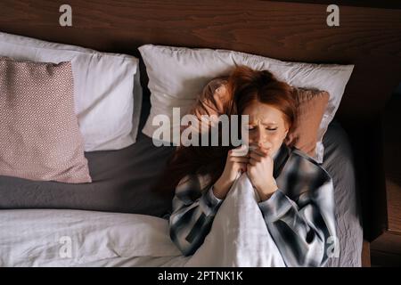 Ein Bild von oben von einer traurigen, depressiven jungen Frau, die sich unter der Decke versteckt und im dunklen Schlafzimmer liegt. Einsam aufgeregte, wunderschöne Frau, die allein liegt Stockfoto