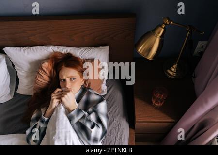 Ein Bild von oben von einer verängstigten jungen rothaarigen Frau, die sich unter einer Decke versteckt und im dunklen Schlafzimmer auf dem Bett liegt. Einsam aufgeregte schöne Frau, die allein im Bett lag Stockfoto