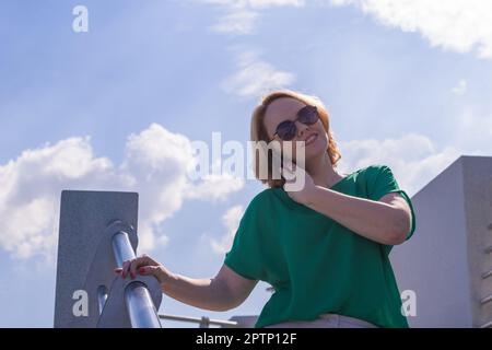 Porträt einer lächelnden Schülerin mit Sonnenbrille, die mit einem Handy am blauen Himmel spricht. Fröhliche Frau, die mit dem Franzosen telefoniert Stockfoto