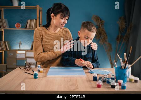 Mom hilft ihrem Sohn, einem Schuljungen, ein Bild mit Farben auf Leinwand zu malen. Gemeinsame Hobbys und Erholung. Stockfoto