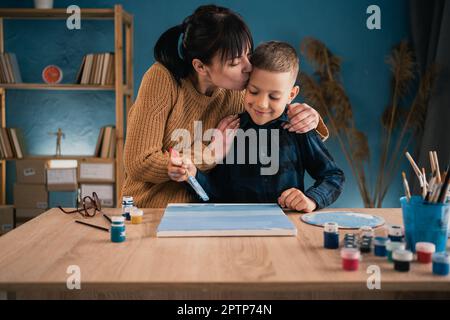 Mom hilft ihrem Sohn, einem Schuljungen, ein Bild mit Farben auf Leinwand zu malen. Eine Frau, die ein Kind umarmt. Gemeinsame Hobbys und Erholung. Stockfoto