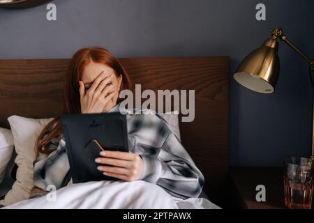 Porträt einer trauernden jungen Frau, die auf dem Bett liegt und das Gesicht mit der Hand bedeckt und weint, während sie den Bilderrahmen hält und das Foto mit Liebe berührt. Stockfoto