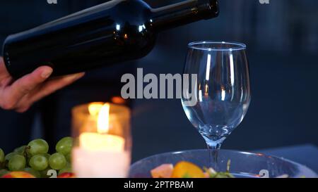 Nahaufnahme eines männlichen Hands mit einer Flasche Wein, der bei einem romantischen Date am Abend ein Getränk serviert. Ein Mann schüttet mit der Hand luxuriösen, teuren Rotwein ins Glas Stockfoto