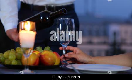 Die Kamera bewegt sich vom Männchen zurück, hält eine Flasche Wein und serviert der Freundin zu einem romantischen Date am Abend ein Getränk. Ein Mann, der Rotwein ins Glas gießt Stockfoto