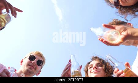 Fröhliche junge Freunde, Männer und Frauen, die Getränke halten und Glasflaschen knallen. Leute auf einer Party, die Spaß haben und trinken. Schließen. Ansicht von unten. Blauer Himmel an Stockfoto