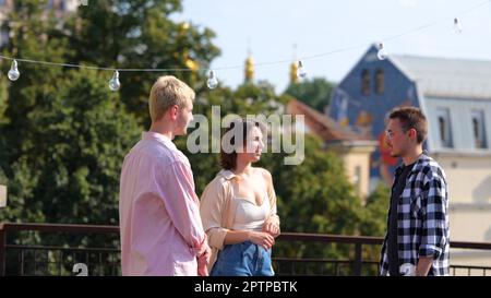 Junge Menschen stehen draußen und plaudern in positiver Stimmung. Kaukasier treffen Freunde auf einem Dach auf einer Party. Sommertag, entspannt. Sie ruht sich draußen aus Stockfoto