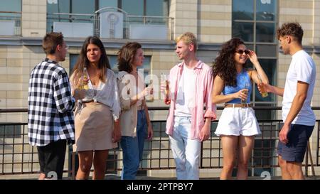 Eine Gruppe junger, ungezwungener Menschen steht draußen mit Drinks auf der Dachparty an sonnigen Tagen, spricht und klappert Flaschen. Glücklicher Weißer, männlich und weiblich Stockfoto