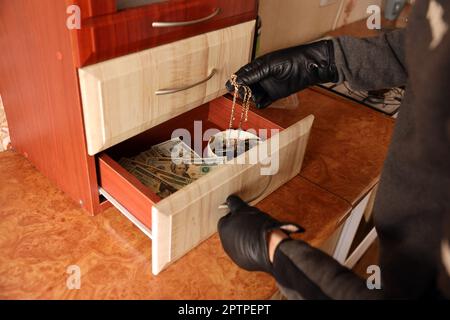 Räuber in schwarzem Outfit und Handschuhe sehen in geöffnetem Regal in der Küche. Der Dieb nimmt die goldene Halskette aus einem Küchenregal Stockfoto