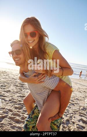 In den Sonnenuntergang reiten. Ein gutaussehender junger Mann, der seiner Freundin am Strand einen Huckepack gibt. Stockfoto