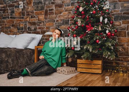 Wunderschöne und positive mutter sitzt auf dem Boden, nachdem sie den Weihnachtsbaum dekoriert hat Stockfoto