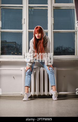 Lässiges Porträt einer jungen Frau in Jeans und Sweatshirt mit Kapuze auf Fensterbank Stockfoto