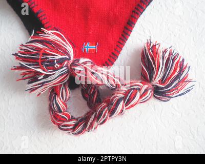 Kappe mit Ohrenklappen im norwegischen Stil. Gestrickter Modeartikel mit Krawatten und Bommeltaschen. Rotes Garn mit schwarz-weißen Fäden. Aufgenähte Minifahne Stockfoto
