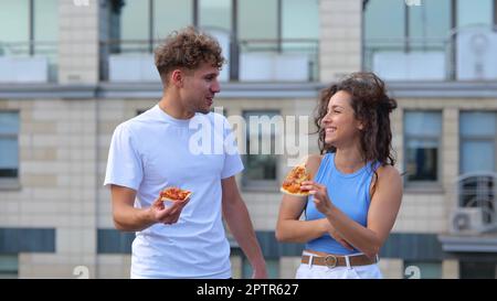 Ein junges weißes Paar, Mann und Frau, die draußen stehen und Pizza auf einer Party auf dem Dach essen. Fröhliche lächelnde Leute, die sich unterhalten und Fastfood probieren Stockfoto