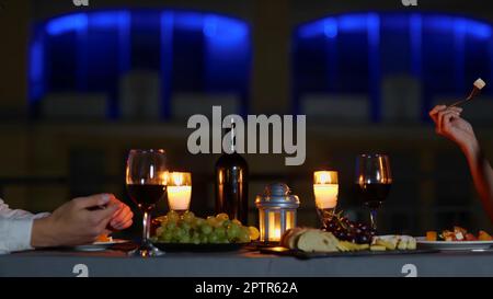 Die Kamera bewegt sich von einem eleganten weißen Paar zurück, das an einem dekorierten Tisch mit Kerzen im Freien sitzt und am Abend ein Date isst. Jubiläumsfeier Stockfoto