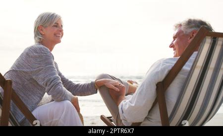 Du liebst den Strand immer noch, genau wie damals. Ein liebevolles Seniorenpaar, das sich an einem Sommertag auf Liegen am Strand entspannt. Stockfoto