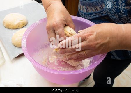 Das Kneten des portugiesischen Süßbrotteigs. Nahaufnahme von Frauenhänden mit Teig in einer Plastikschüssel, die auf dem Küchentisch geformt wird Stockfoto