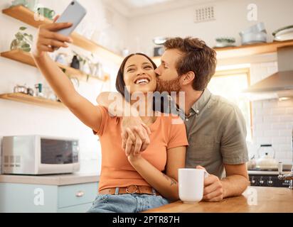 Ein junges, fröhliches, gemischtrassiges Paar, das zu Hause ein Selfie mit einem Telefon macht. Fröhlicher, weißer Freund, der seine gemischte Freundin auf der C küsst Stockfoto