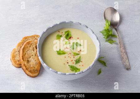 Cremefarbene Sellerie-Suppe mit Sahne, garniert mit Sellerieblättern, pinkfarbenem Pfeffer und serviert mit knusprigem Toast auf hellgrauem Hintergrund, Draufsicht Stockfoto