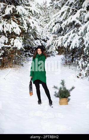 Ein junges, hübsches Mädchen mit grünem Pullover und Hut steht im Winterwald mit einem Korb mit Pinienästen und einer Schnur-Tüte mit Mandarinen. Stockfoto