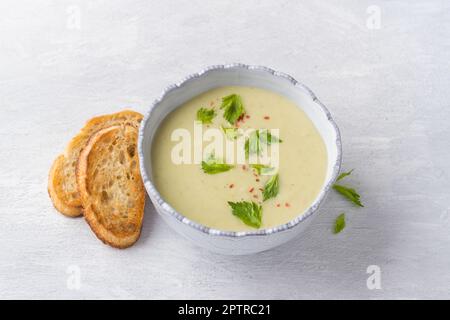 Cremefarbene Sellerie-Suppe mit Sahne, garniert mit Sellerieblättern, pinkfarbenem Pfeffer und serviert mit knusprigem Toast auf hellgrauem Hintergrund, Draufsicht Stockfoto