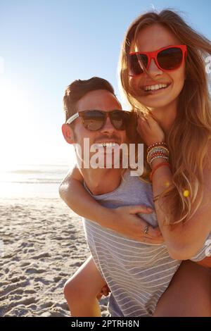 Genießen Sie einen perfekten Tag unter der Sonne. Porträt eines gutaussehenden jungen Mannes, der seiner Freundin am Strand einen Huckepack gibt. Stockfoto