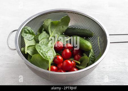 Frisch gewaschene Spinatblätter, Kirschtomaten und Gurken in einem Sieb auf hellgrauem Hintergrund. Zubereitung eines gesunden hausgemachten Salats Stockfoto