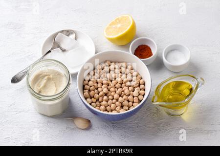 Zutaten für die Herstellung von libanesischem Hummus: Trockene Kichererbsen, Tahini-Sesampaste, Olivenöl, Knoblauch, Zitrone, Eis, Paprika und Salz auf hellgrauem Hintergrund, Stockfoto
