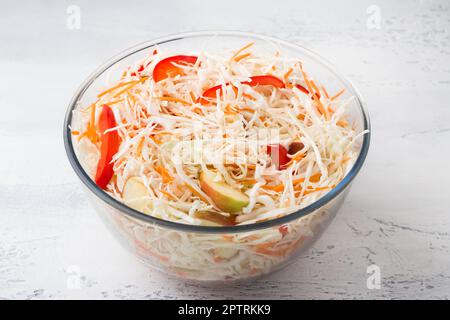 Glasschüssel mit gehacktem Kohl mit geriebenen Karotten, Äpfeln und Gemüsepaprika auf hellgrauem Hintergrund. Köstliche hausgemachte Speisen Stockfoto