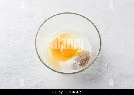 Köstlicher Grießkuchen: Saure Sahne oder Joghurt, zwei Eier, Zucker, Vanillezucker und eine Prise Salz in einer Glasschüssel auf hellgrauem Hintergrund, t Stockfoto