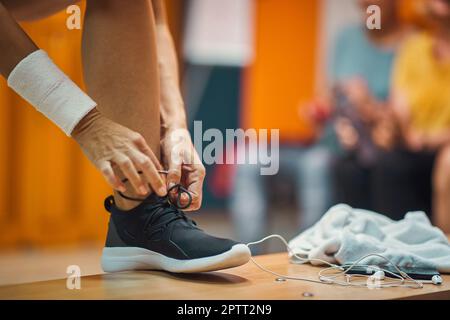 Junge Frau bindet ihren Schuh im Umkleideraum des Fitnessstudios, mit Kopfhörern und Handtuch auf der Bank, macht sich bereit für das Training. Helath, Lifestyle, aktives Konzept. Stockfoto