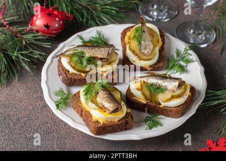 Hausgemachte warme Sandwiches mit Sprotten, Ei, Käse und eingelegter Gurke auf rundem Teller mit braunem Hintergrund Stockfoto