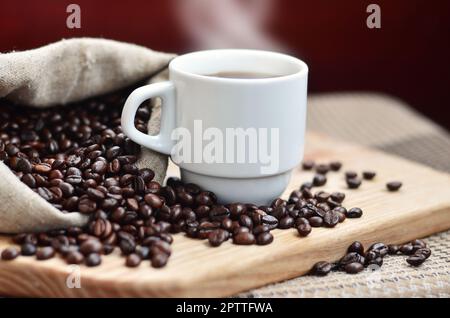 Einen vollen Beutel der braunen Bohnen und eine weiße Tasse heißen Kaffee liegt auf einem Holz- Oberfläche. Attribute, die mit der Zubereitung von Kaffee im Zusammenhang Stockfoto