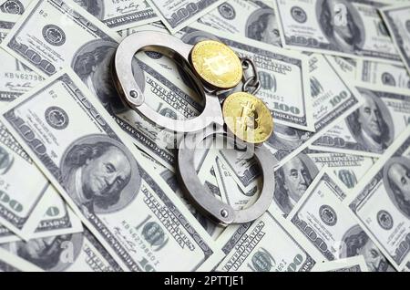 Polizei Handschellen und Bitcoins liegen auf eine große Anzahl von Dollarnoten. Das Konzept der Probleme mit dem Gesetz während der Illegalen cryptocurrency Bergbau- und Stockfoto