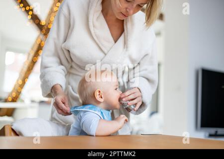 Freizeitkonzept für häusliche Momente. Säuglingsjunge trinkt Wasser aus kleinem Glas mit Hilfe seiner Mutter im kuscheligen Bademantel Stockfoto