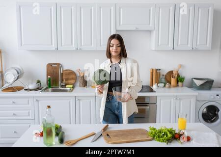 Brünette Frau mit Brokkoli und Schüssel in der Nähe von Gemüse in der Küche, Stockbild Stockfoto