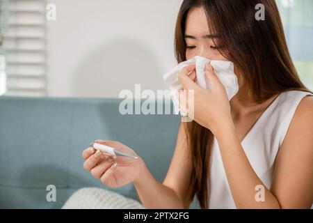 Kranke Frau haben Symptom krank hohes Fieber sie prüft ihre Temperatur mit Thermometer im Wohnzimmer zu Hause, junge Frau leidet an Grippe Erkältung und Stockfoto