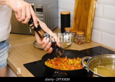 Nahaufnahme der Zugabe von schwarzem Pfeffer in Gemüse in einer Pfanne. Frau benutzt Pfeffermühle in der Küche. Stockfoto