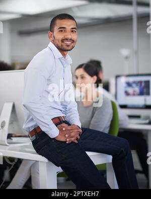 Hes hat sich Geschäft angesammelt. Ein hübscher Geschäftsmann sitzt auf seinem Schreibtisch mit ein paar Kollegen im Hintergrund Stockfoto