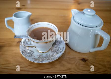 Eine weiße porzellan-Teetasse und eine dekorierte Untertasse auf einem Holztisch mit einer Teekanne und einem Milchkännchen. Stockfoto