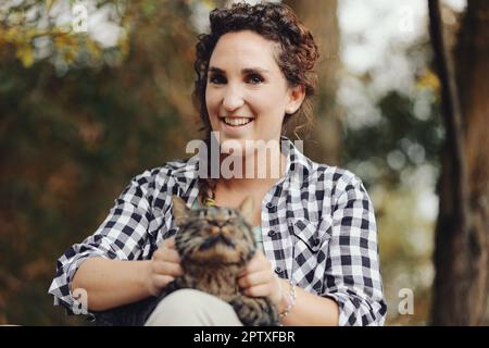 In einem öffentlichen Park streichelt und kuschelt eine Frau in einem schwarz-weißen Karohemd mit einer großen Katze, die sie gefunden hat. Er genießt das Vergnügen, weil Katzen wissen, wie es geht Stockfoto