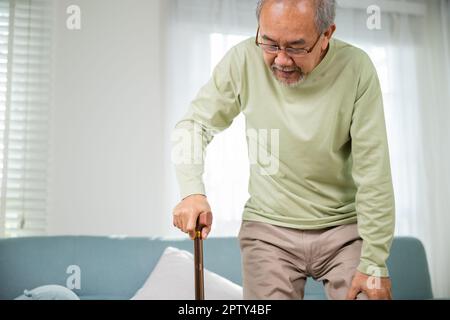 Asiatischer alter Mann mit einer Brille, die mit Gehstock vom Sofa aus aufsteht, ältere Menschen mit Knieschmerzen, die den Griff des Rohrs halten, Senior DIS Stockfoto