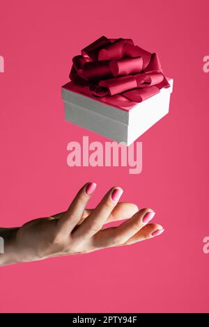 Weibliche Hand wirft in die Luft oder fängt weiße Geschenkbox mit Schleife auf dem Hintergrund von Color of the Year 2023 Viva Magenta. Schwebende Gabe. Vertikal Stockfoto