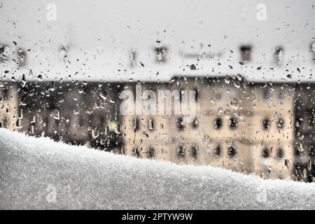 Blick vom Fenster auf das Winterauftauen. Schneeverwehungen und Tropfen auf dem Glas schmelzen. Mehrstöckiges Gebäude mit vielen Fenstern. Weihnachten Neujahr Postkutsche Stockfoto