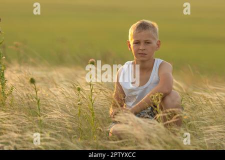 Ein süßer Junge in einem weißen T-Shirt, der auf einem Hügel zwischen Federgras sitzt. Sommerzeit und fröhliches Kindheitskonzept. Stockfoto