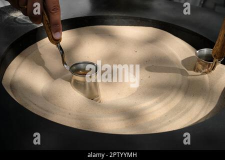 Nahaufnahme der türkischen Kaffeezubereitung in Cezve auf Sand in der Kaffeebar. Ein professioneller Barista bereitet Kaffee manuell auf traditionelle Weise und mit modernem Material zu Stockfoto